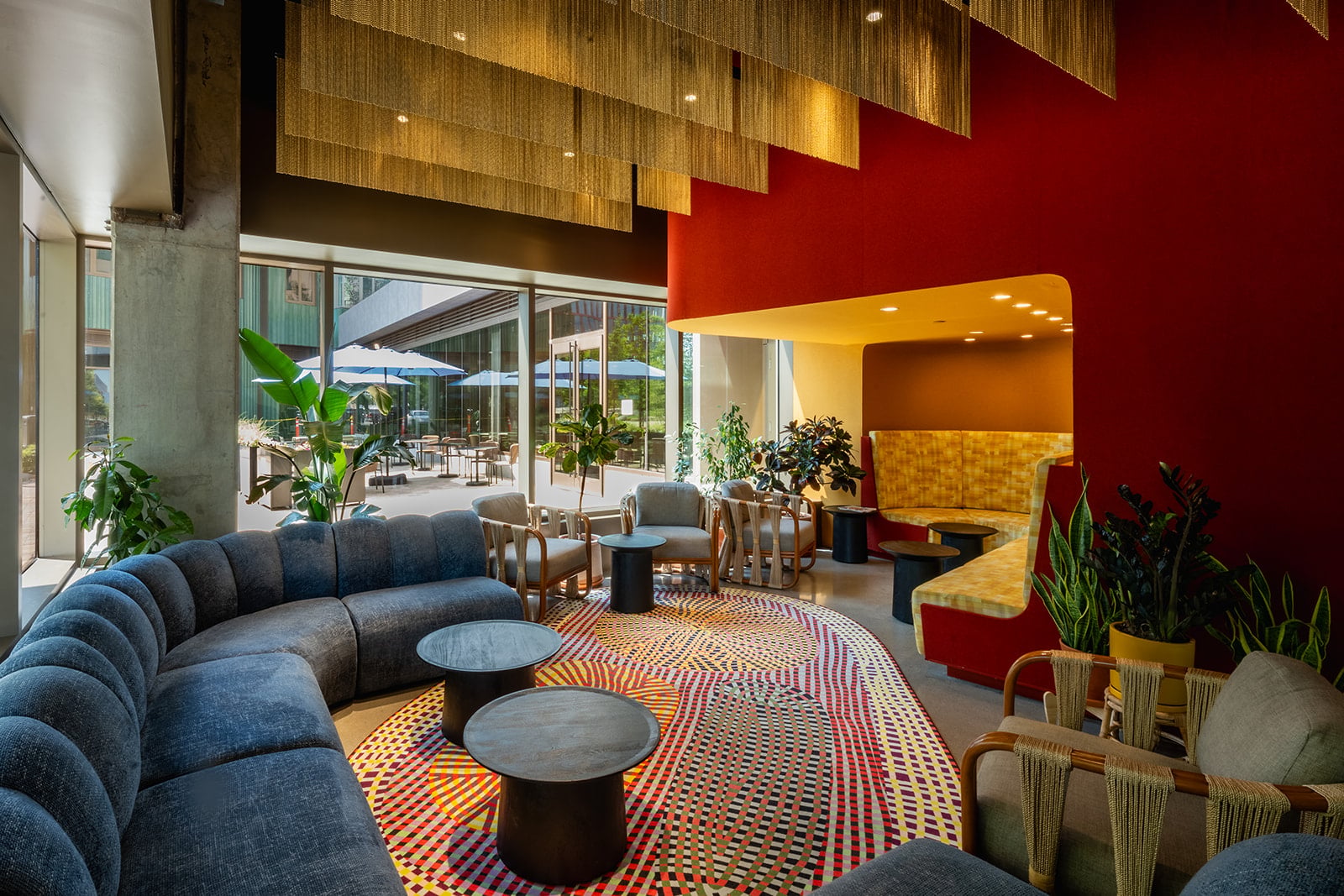 A modern hotel lounge with a curved blue sofa, colorful patterned rug, and seating areas by large glass walls overlooking outdoor tables with umbrellas.