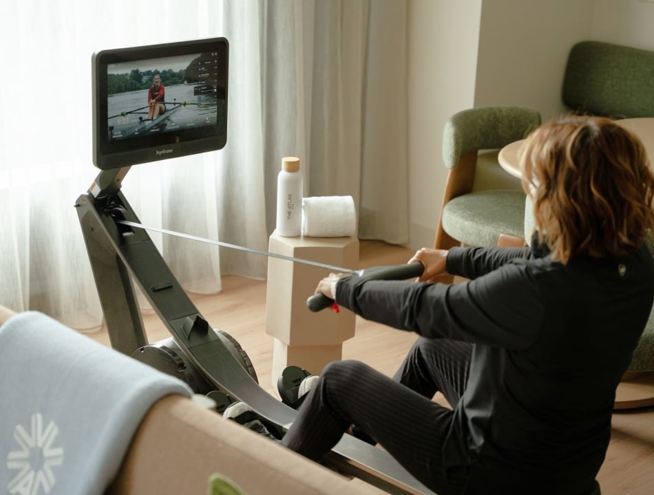Person in a black outfit sits on a Hydrow rowing machine, watching the screen showing a person rowing on water; a bottle and towel sit on a nearby stand in a living room.