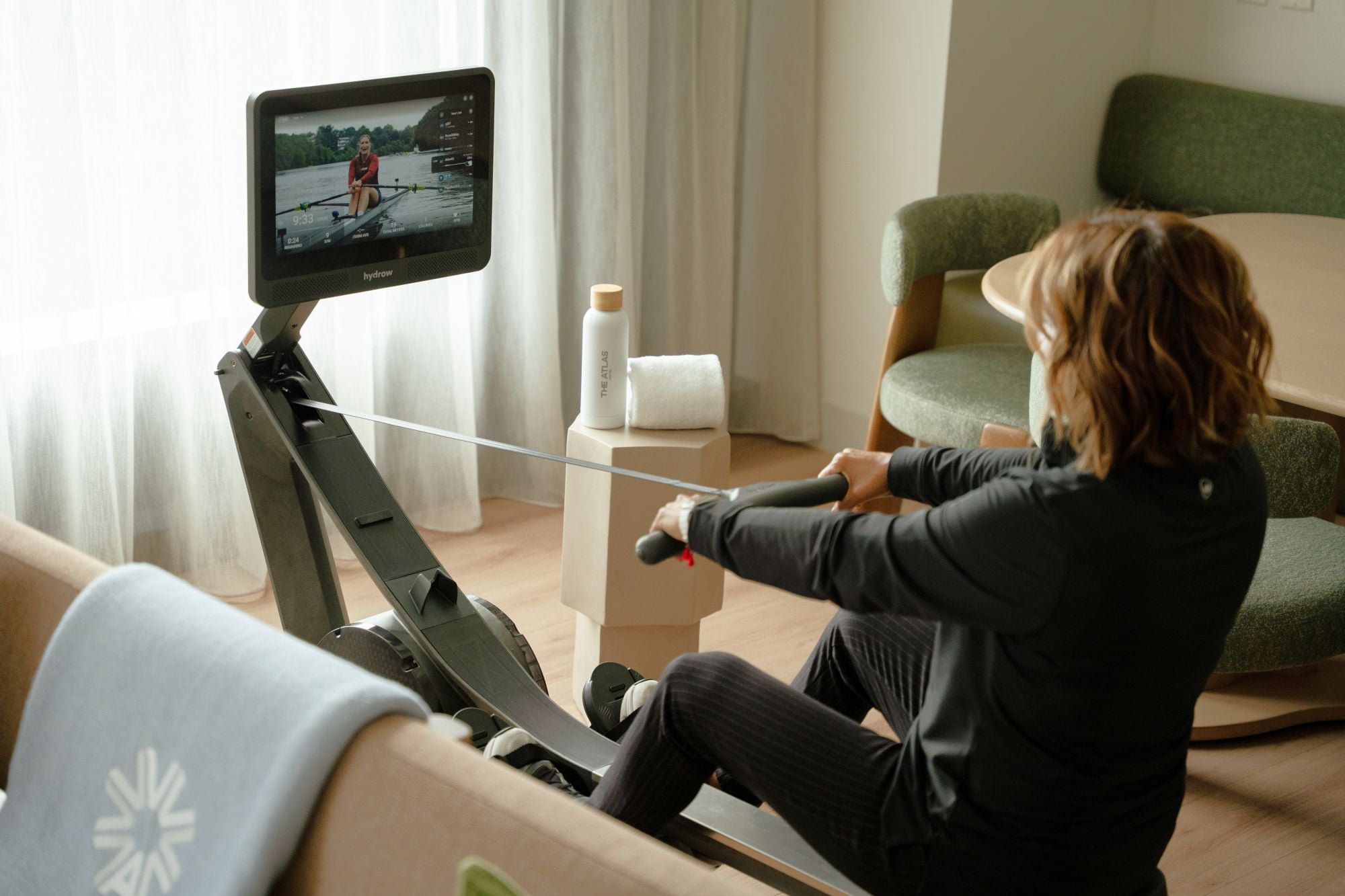 Person in a black outfit sits on a Hydrow rowing machine, watching the screen showing a person rowing on water; a bottle and towel sit on a nearby stand in a living room.