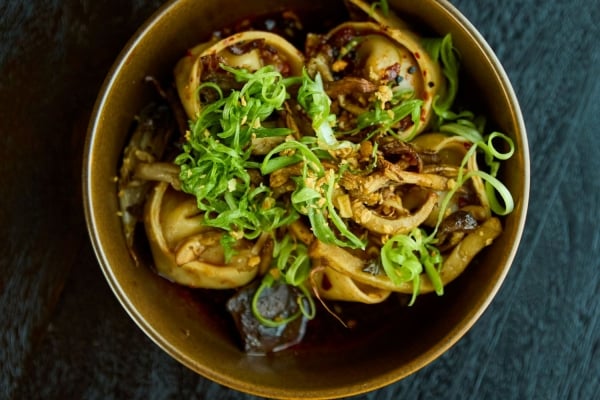 Brown ceramic bowl of noodles topped with green onions, mushrooms, and crispy fried bits on a dark wooden surface