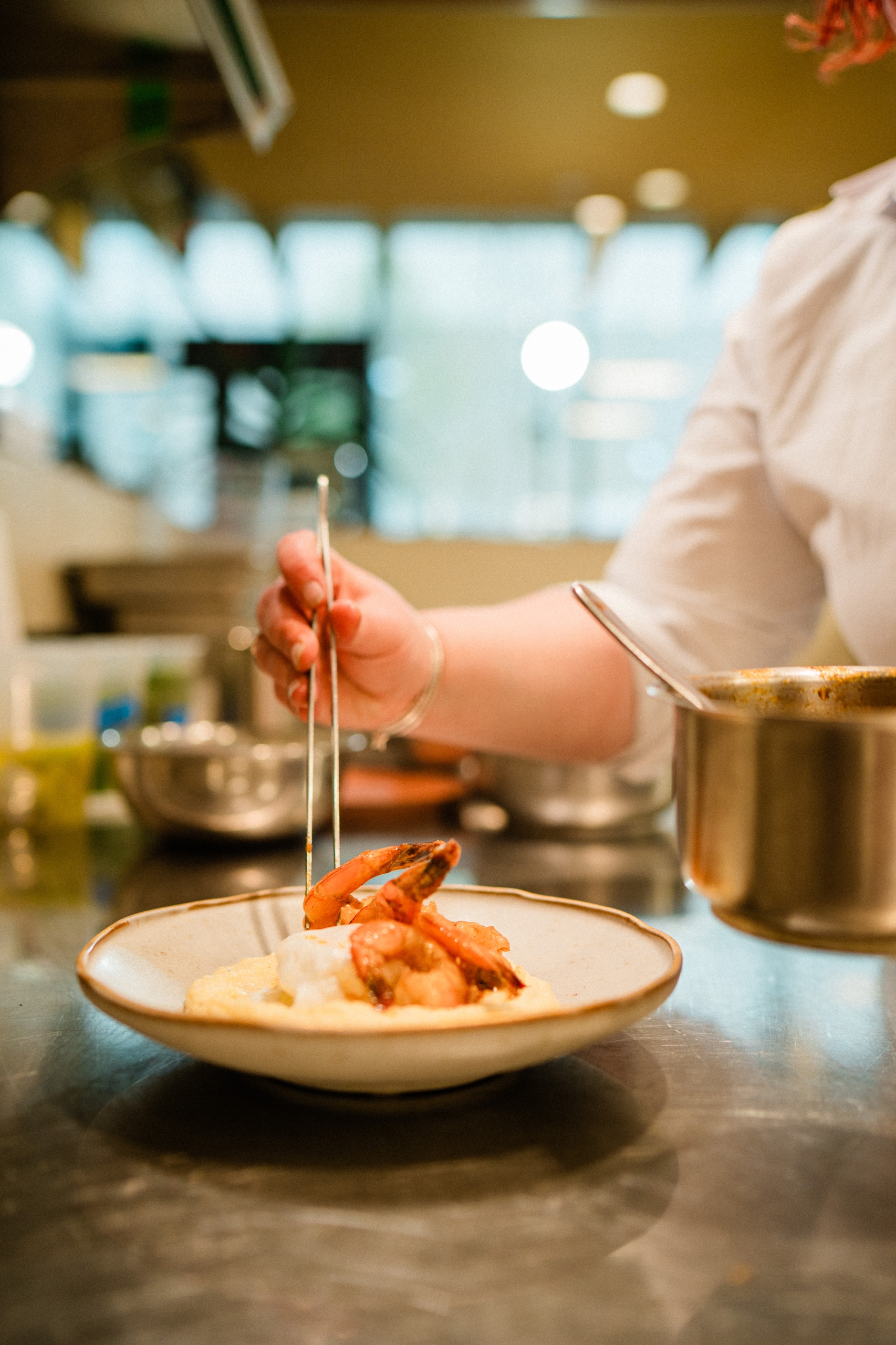 A cook uses tongs to plate shrimp and sauce on a white plate in a stainless steel kitchen.