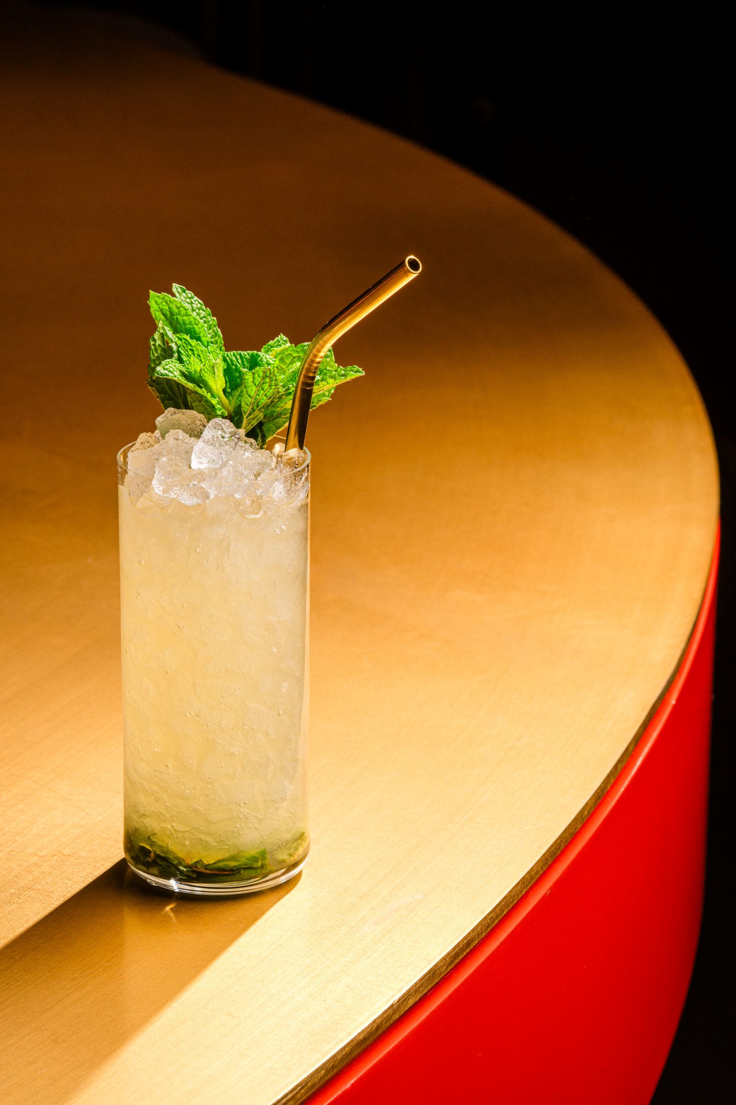 Tall glass cocktail with crushed ice and fresh mint, placed on a warm gold bar surface with a metal straw.