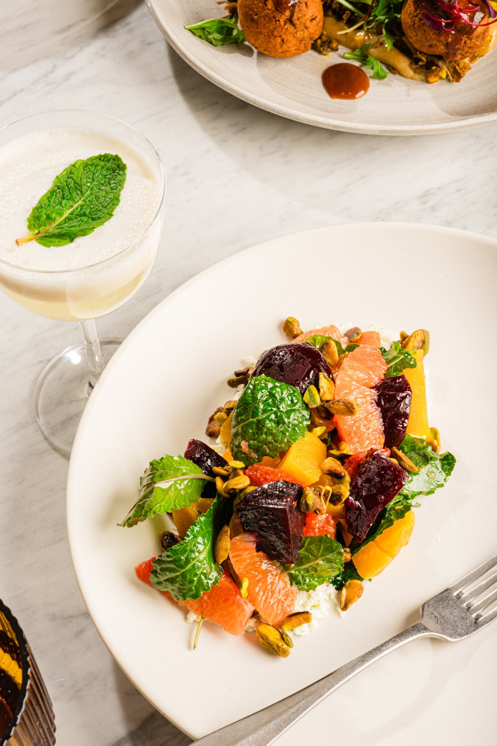 White plate holds citrus salad with orange segments, beets, greens and pistachios on a marble table, with a mint-leaf drink nearby.
