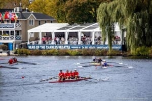Four rowers in a red four-oared boat race on the Charles River during the Head of the Charles Regatta, with a white tented spectator area and onlookers in the background.