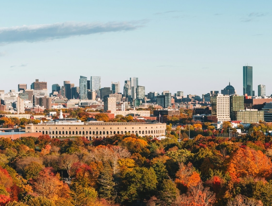 City skyline rising above a vibrant autumn forest with red, orange, and yellow trees in the foreground.