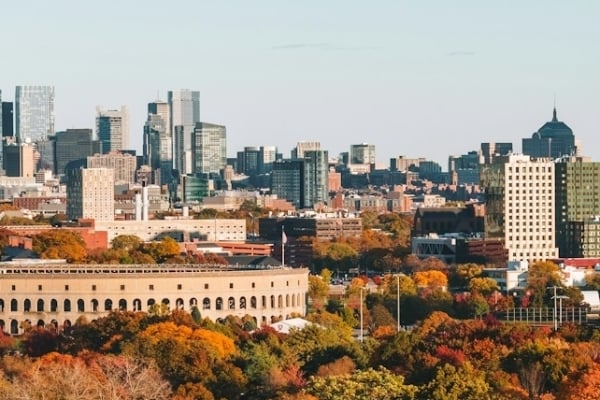 City skyline rising above a vibrant autumn forest with red, orange, and yellow trees in the foreground.