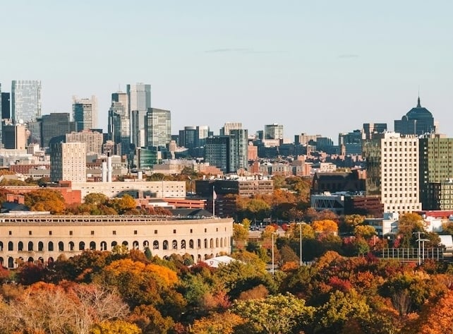 City skyline rising above a vibrant autumn forest with red, orange, and yellow trees in the foreground.
