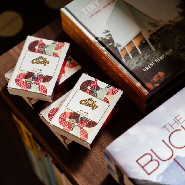 Several boxes of 'the coop' playing cards with colorful swirl patterns on a wooden table, beside a book titled 'The Bucket List'.
