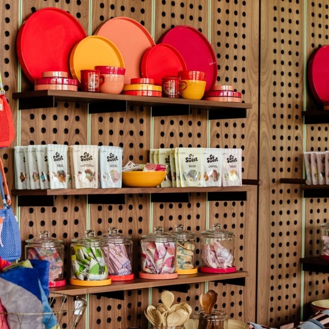 Colorful kitchenware display on wooden shelves: plates, bowls, mugs, and jars in red, pink, orange, and yellow.