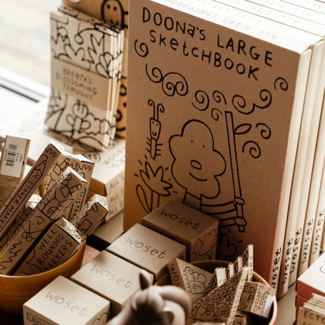 Brown sketchbook titled 'Doona's Large Sketchbook' with doodle drawings, surrounded by smaller boxed items in a display case.