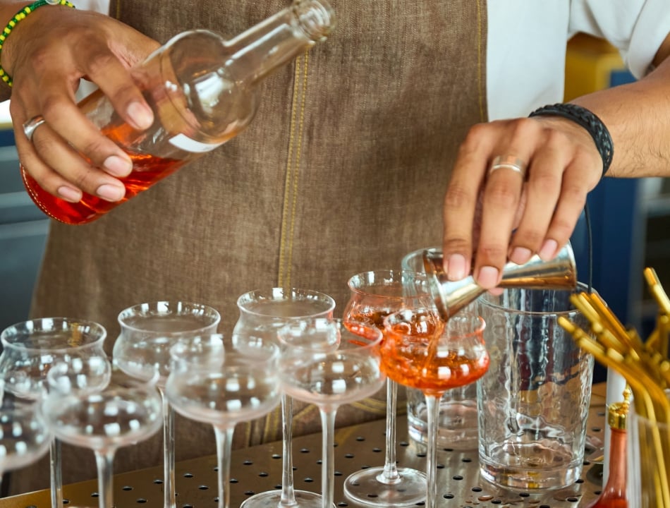 Bartender pours red liquor from a bottle into stemmed glasses lined up on a bar, with a brown apron visible in the background.
