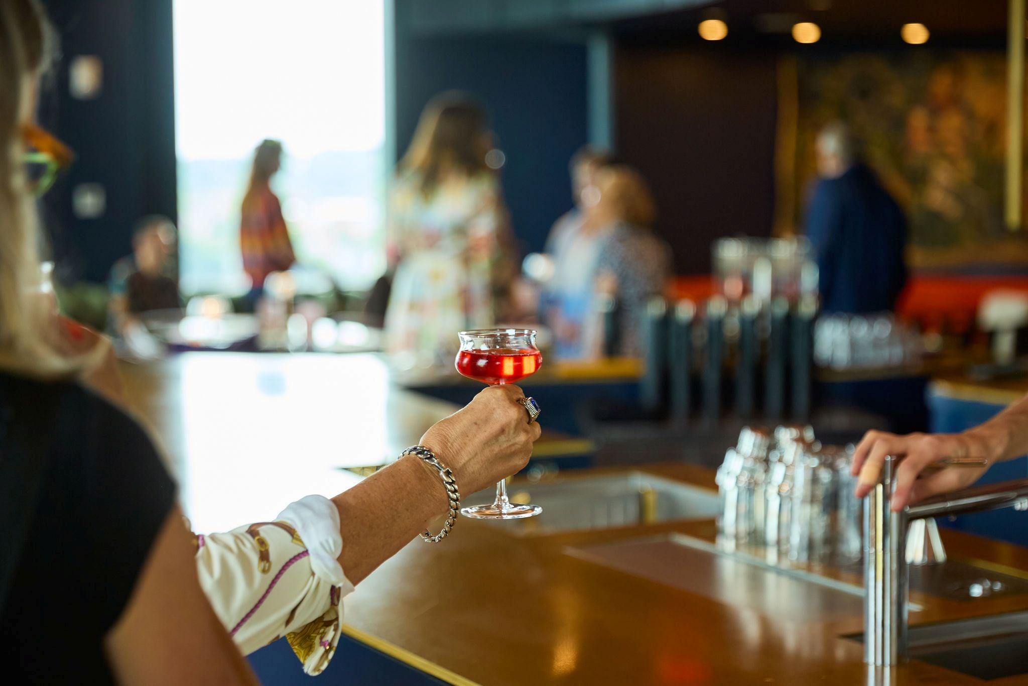 A hand with a bracelet and ring holds a small red cocktail in a stemmed glass at a bar, with blurred patrons in the background.
