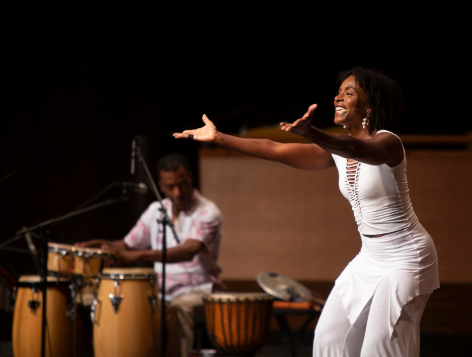A woman in a white outfit extends her arms while smiling on stage, wearing a headset mic as a guitarist/percussionist plays in the background.