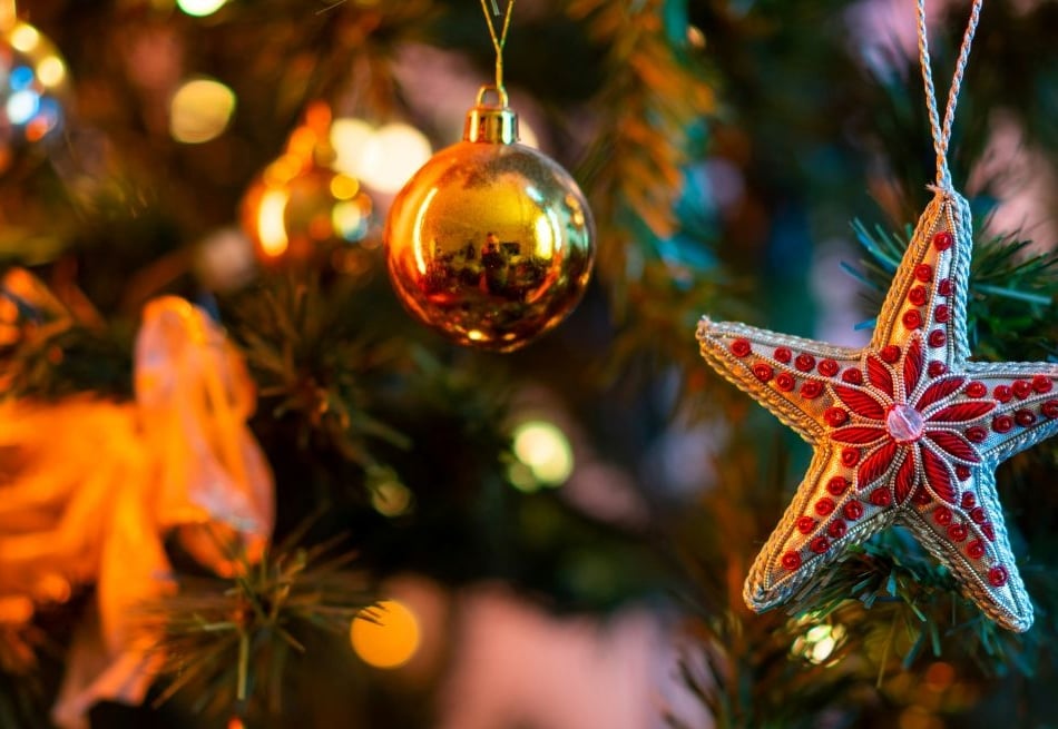Close-up of a beaded red star ornament hanging on a Christmas tree with gold baubles and warm string lights.