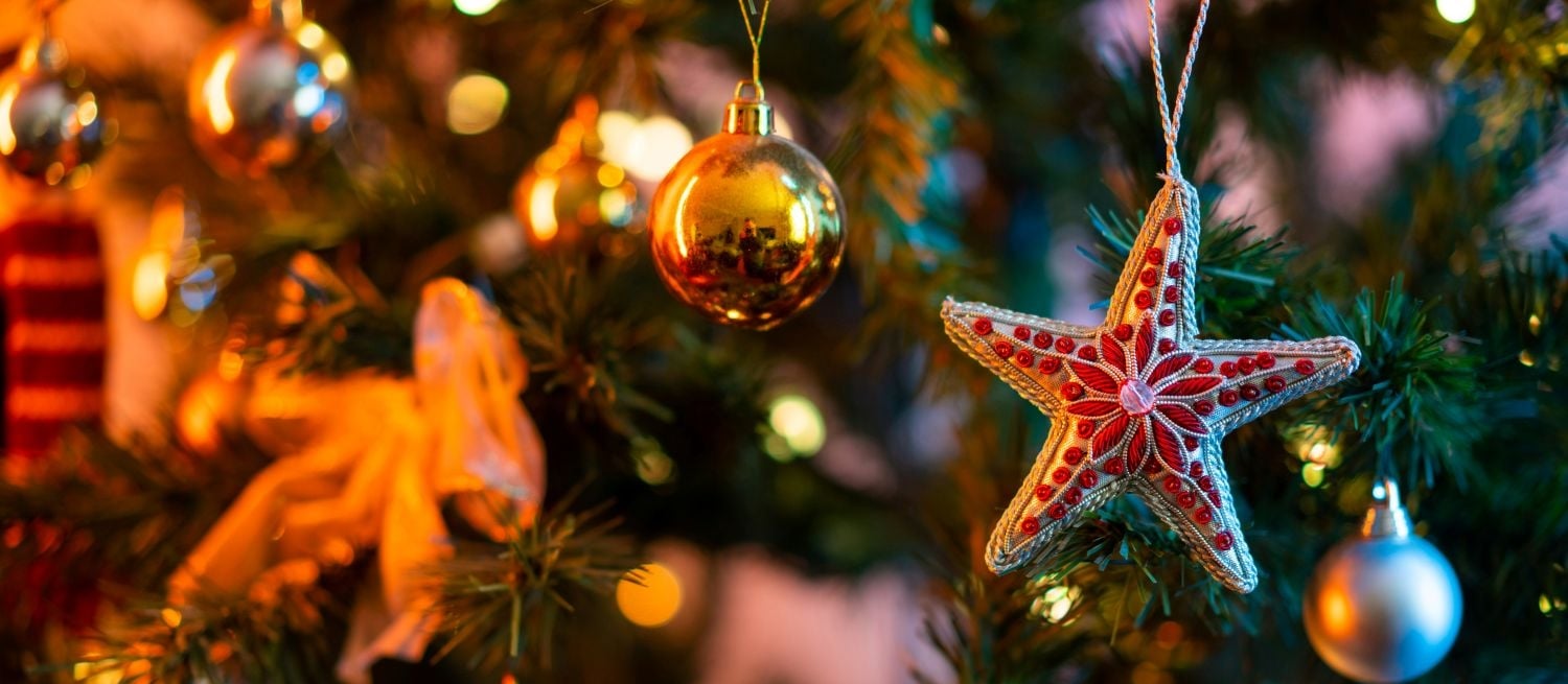 Close-up of a beaded red star ornament hanging on a Christmas tree with gold baubles and warm string lights.