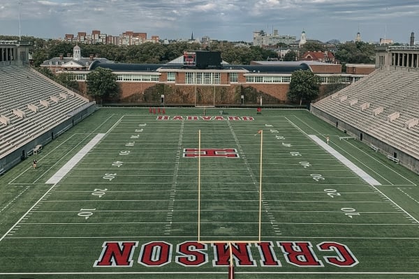 Overhead view of a college football stadium with empty concrete stands and a green field, goalposts, and a distant city skyline.