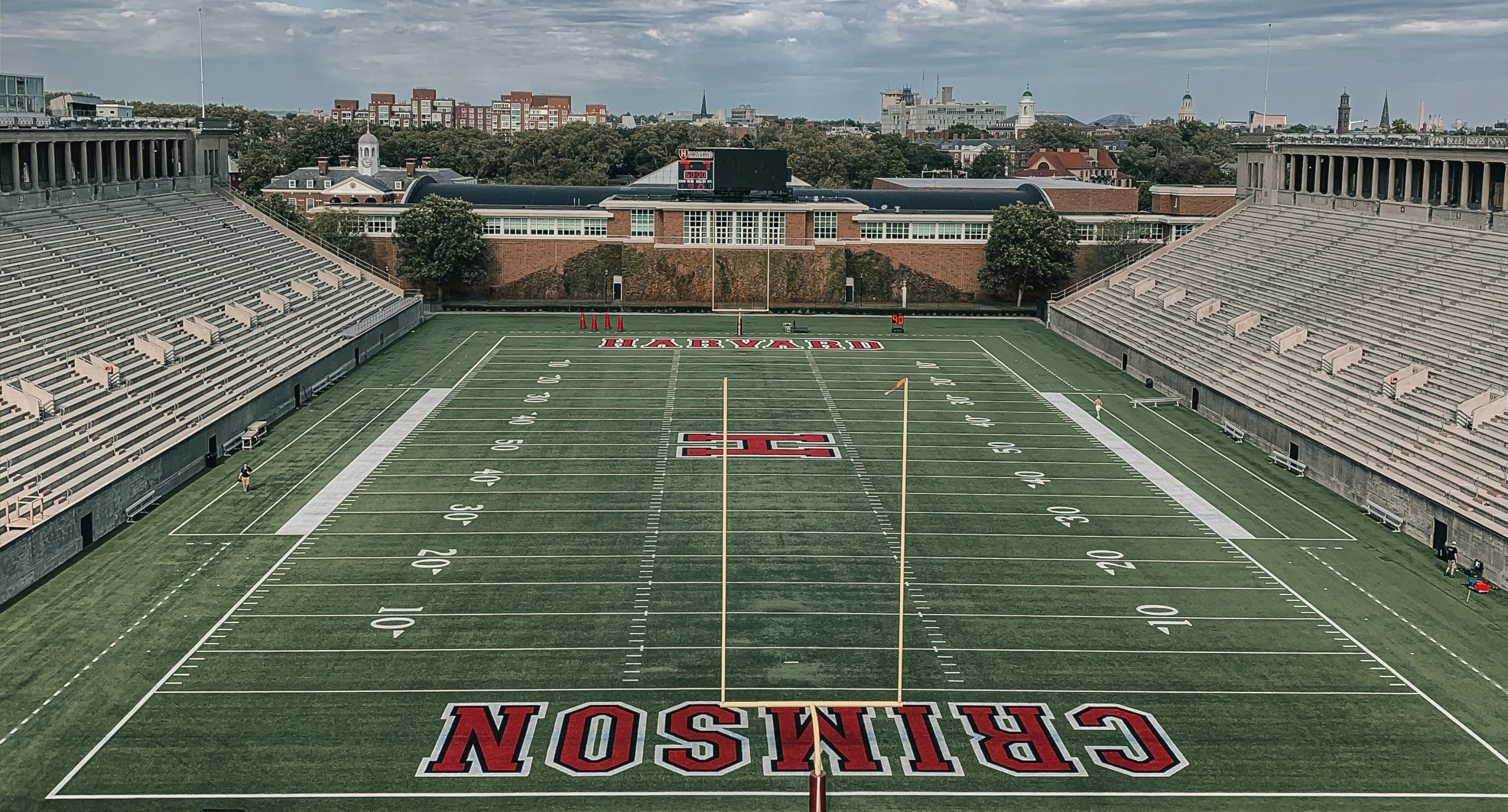 Overhead view of a college football stadium with empty concrete stands and a green field, goalposts, and a distant city skyline.